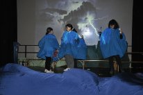 Die Schüler*innen stellen auf dem Bild ein Gewitter dar. Sie tragen blaue Regenmäntel. Im Hintergrund ist ein Bild eines Blitzes zu erkennen.